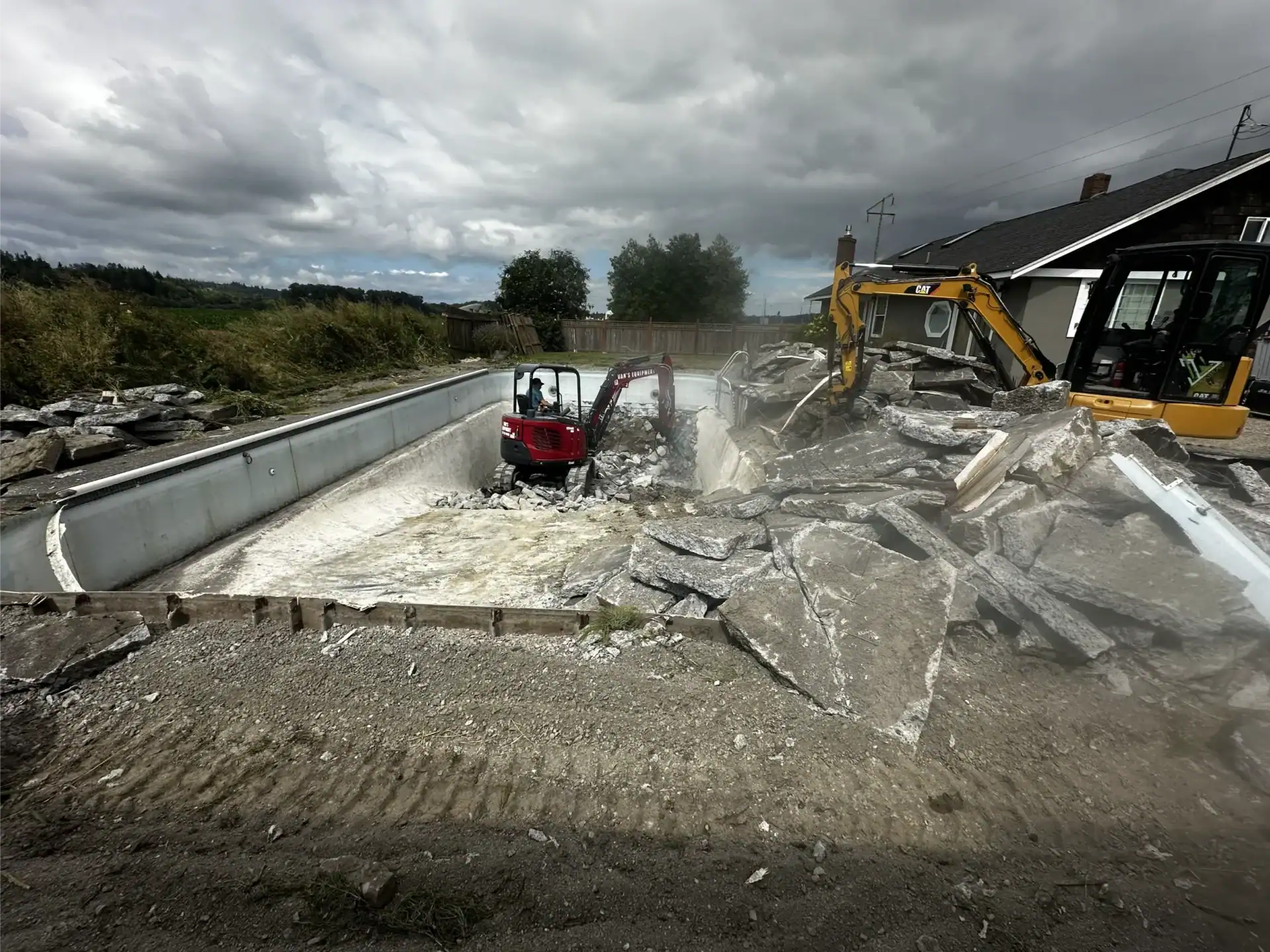 Two excavators demolish a concrete swimming pool, breaking up and removing large pieces of debris