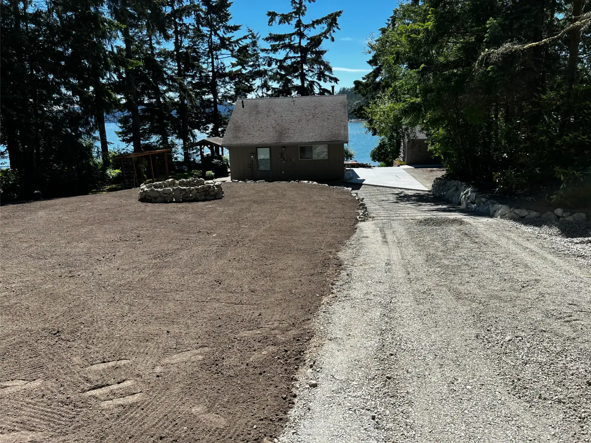 a gravel driveway to a small cottage and a cleared dirt area with a circular stone fire pit