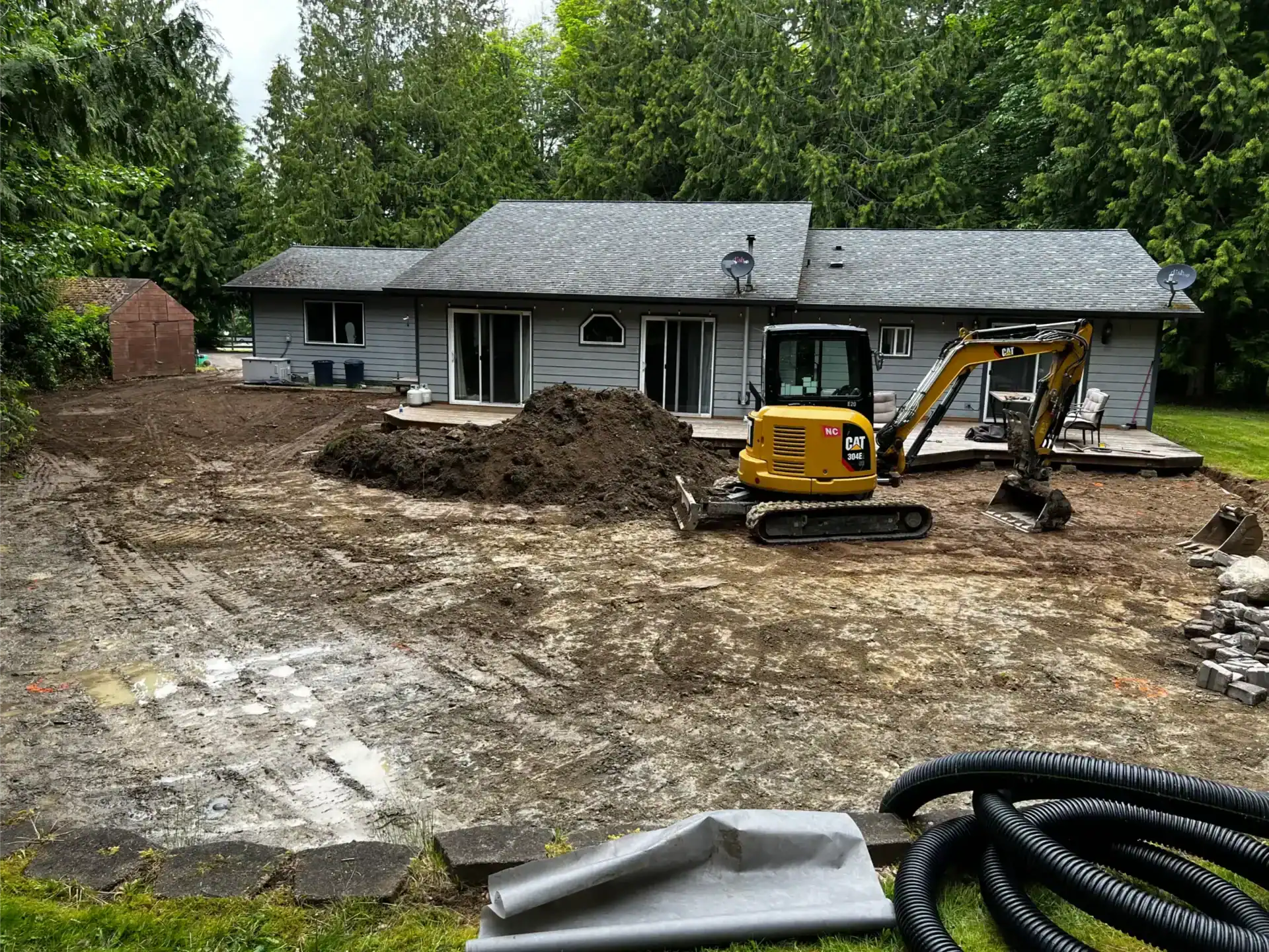 A small excavator sits on dirt behind a gray house, with piles of soil and black drainage pipes nearby, showing ongoing excavation and grading work